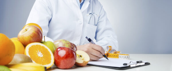 Professional nutritionist working at desk and writing medical records with fresh fruit on foreground